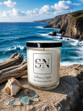 A 9 oz candle by ‘Signature Note Candle Co’ in a clear glass jar with a black lid, labeled 'Azure Cliffs' sits on a rocky beach with ocean waves and cliffs in the background.