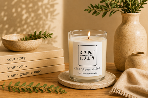 A lit candle in a clear glass jar sits on a round coaster on a wooden table, surrounded by books, a ceramic bowl, a vase with greenery, and a folded knit cloth. The candle label reads 'Signature Note Candle Co' and 'Black Raspberry Vanilla'. 