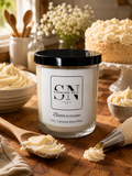 A candle jar labeled 'Signature Note Candle Co. Buttercream' sits on a wooden table surrounded by frosting bowls, a piping bag, and a decorated cake in a warm kitchen-like setting.