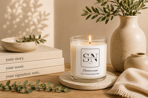 A lit candle in a clear glass jar sits on a round coaster on a wooden table, surrounded by books, a ceramic bowl, a vase with greenery, and a folded knit cloth. The candle label reads 'Signature Note Candle Co' and 'Buttercream'.