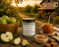 A 7 oz candle by ‘Signature Note Candle Co’ in a clear glass jar with a black lid, labeled 'Caramel Apple' styled in an orchard on a wooden table with a candle, a basket of green apples, a bowl of brown sugar, a caramel apple on a plate, caramels, and a barn in the background. 