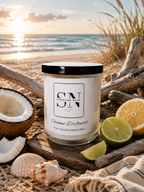 A 9 oz candle by ‘Signature Note Candle Co’ in a clear glass jar with a black lid, labeled 'Coconut Driftwood' styled on a beach with driftwood, seashells, a coconut, and limes.
