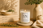 A lit candle in a clear glass jar sits on a round coaster on a wooden table, surrounded by books, a ceramic bowl, a vase with greenery, and a folded knit cloth. The candle label reads 'Signature Note Candle Co' and 'Coconut Driftwood'.