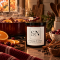 A 9 oz candle jar labeled 'Signature Note' 'Crimson & Clove' wax, styled on a rustic wooden table with oranges, cranberries, cinnamon sticks, and a cozy simmer pot in the background.