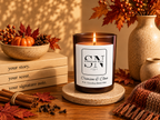 A lit amber glass candle with a white label reading 'Signature Note' and 'Crimson & Clove' sits on a stone coaster on a wooden table, styled with fall leaves, cinnamon sticks, books, a small pumpkin, and ceramic vases.
