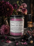 A 9 oz candle by ‘Signature Note Candle Co’ in a clear glass jar with a black lid, labeled 'Dahlias In Obsidian' sits in a dark, moody room with deep purple dahlias, obsidian, and candles.