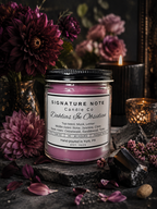 A 9 oz candle by ‘Signature Note Candle Co’ in a clear glass jar with a black lid, labeled 'Dahlias In Obsidian' sits in a dark, moody room with deep purple dahlias, obsidian, and candles.