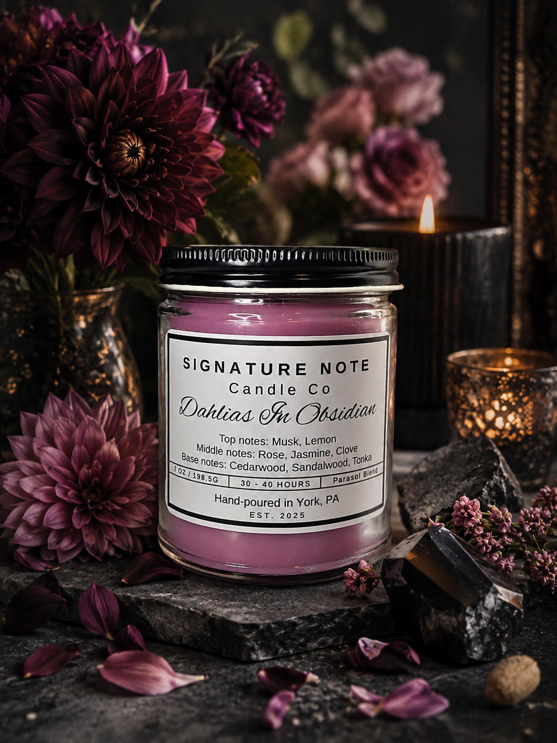 A 9 oz candle by ‘Signature Note Candle Co’ in a clear glass jar with a black lid, labeled 'Dahlias In Obsidian' sits in a dark, moody room with deep purple dahlias, obsidian, and candles.