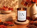 A lit candle in a dark amber glass jar sits on a round coaster on a wooden table, surrounded by fall-themed props including books, cinnamon sticks, berries, a small pumpkin, and a vase with autumn leaves. The candle label reads 'Signature Note' and 'Honey Spiced Pear'.