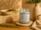 A lit candle in a clear glass jar sits on a round coaster on a wooden table, surrounded by books, a ceramic bowl, a vase with greenery, and a folded knit cloth. The candle label reads 'Signature Note Candle Co' and 'Lakeside Retreat'. 