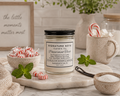 A 7 oz candle by ‘Signature Note Candle Co’ in a clear glass jar with a black lid, labeled 'Peppermint Twist' styled in a kitchen on a marble table with a candle, a plate of waffles, a bowl of peppermints, a bowl of sugar, mint leaves, vanilla beans and a cup with a candy cane and sugar cubes in the background. 