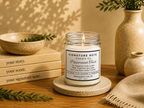 A lit candle in a clear glass jar with cream wax sits on a round coaster on a wooden table, surrounded by books, a ceramic bowl, a vase with greenery, and a folded knit cloth. The candle label reads 'Signature Note Candle Co' and 'Peppermint Twist'. 