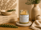 A lit candle in a clear glass jar with orange wax sits on a round coaster on a wooden table, surrounded by books, a ceramic bowl, a vase with greenery, and a folded knit cloth. The candle label reads 'Signature Note Candle Co' and 'Pumpkin Pecan Waffles'. 
