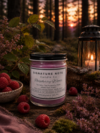 A 9 oz candle by ‘Signature Note Candle Co’ in a clear glass jar with a black lid, labeled 'Raspberry Woods' sits outside in the woods with raspberries, florals, and a lantern.