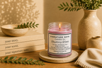 A lit candle in a clear glass jar sits on a round coaster on a wooden table, surrounded by books, a ceramic bowl, a vase with greenery, and a folded knit cloth. The candle label reads 'Signature Note Candle Co' and 'Raspberry Woods'.