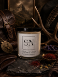 A 9 oz candle by ‘Signature Note Candle Co’ in a clear glass jar with a black lid, labeled 'Saffron & Stone' styled on a dark stone surface with leather, antlers, spices, and a compass in a moody rustic setting.