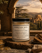 A 9 oz candle by ‘Signature Note Candle Co’ in a clear glass jar with a black lid, labeled 'Sandalwood & Cedar' styled on a stone on a table with beads, cedarwood, and a stone vase with greenery. A window shows an ancient temple and hillsides in the background.