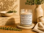 A lit candle in a clear glass jar sits on a round coaster on a wooden table, surrounded by books, a ceramic bowl, a vase with greenery, and a folded knit cloth. The candle label reads 'Signature Note Candle Co' and 'Sandalwood & Cedar'. 
