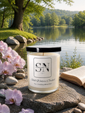A 9 oz candle by ‘Signature Note Candle Co’ in a clear glass jar with a black lid, labeled 'Still Waters Orchid' sits on a stone beside a lake, with pink orchids, rocks, trees, and an open book in the background.