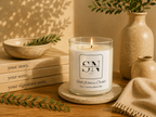 A lit candle in a clear glass jar sits on a round coaster on a wooden table, surrounded by books, a ceramic bowl, a vase with greenery, and a folded knit cloth. The candle label reads 'Signature Note Candle Co' and 'Still Waters Orchid'.