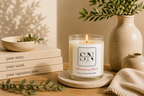 A lit candle in a clear glass jar sits on a round coaster on a wooden table, surrounded by books, a ceramic bowl, a vase with greenery, and a folded knit cloth. The candle label reads 'Signature Note Candle Co' and 'Strawberry Basil'.
