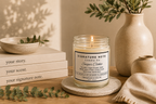 A lit candle in a clear glass jar sits on a round coaster on a wooden table, surrounded by books, a ceramic bowl, a vase with greenery, and a folded knit cloth. The candle label reads 'Signature Note Candle Co' and 'Sugar Cookie'.
