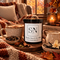 A jar candle labeled 'Vanilla Chai & Oat Milk' by Signature Note Candle Co. is styled on a rustic wooden table with fall-themed props including a mug of coffee, a pitcher of milk, books, a candle, a chair with a blanket and pillow, in a cozy indoor setting.