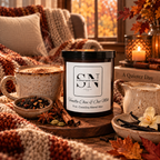 A jar candle labeled 'Vanilla Chai & Oat Milk' by Signature Note Candle Co. is styled on a rustic wooden table with fall-themed props including a mug of coffee, a pitcher of milk, books, a candle, a chair with a blanket and pillow, in a cozy indoor setting.