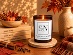 A lit amber glass candle in a speckled beige coaster holder, styled on a wooden table with fall decor including books, cinnamon sticks, red leaves, pinecones, a small pumpkin, and ceramic vases. The candle label reads Signature Note and Vanilla Chai & Oat Milk.